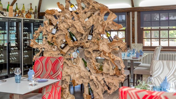 L’intérieur d’un restaurant présente un décor original, avec une pièce maîtresse en bois abritant des bouteilles de vin, des sièges modernes et une vue dégagée grâce à de grandes fenêtres.