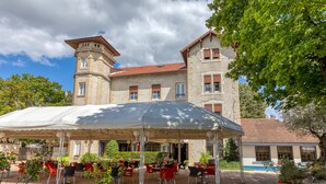 Une cour avec des sièges en plein air, un espace couvert et un bâtiment doté d’une tour et de nombreuses fenêtres.