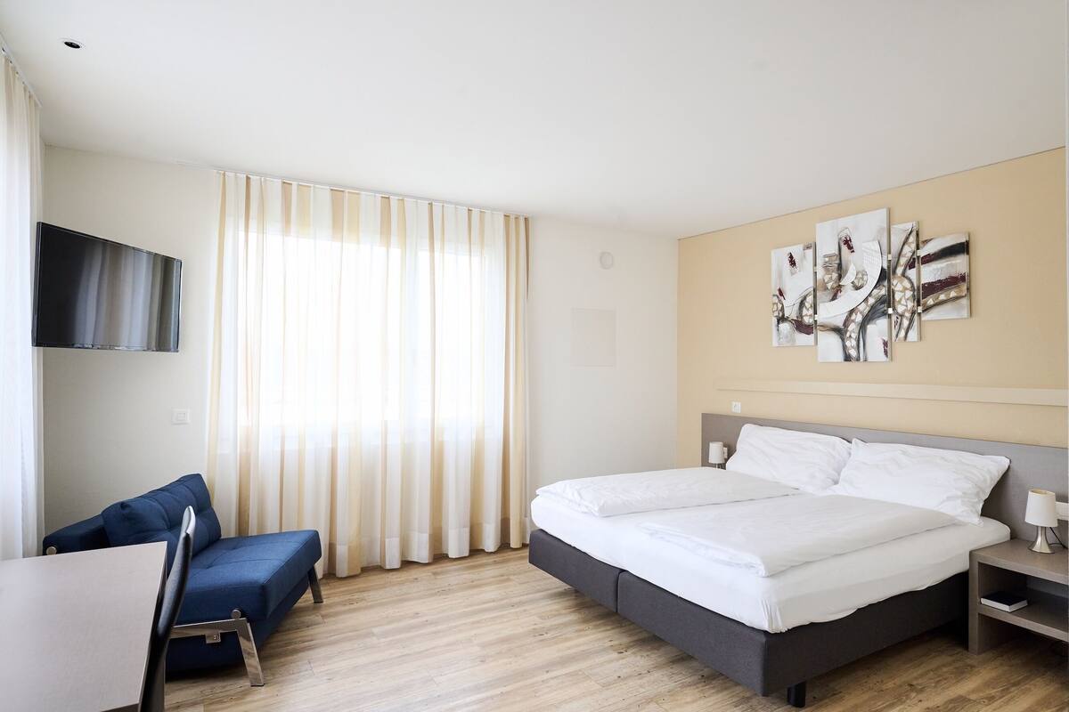 A modern hotel room with a large bed, a sofa, a TV, and artwork on the wall.