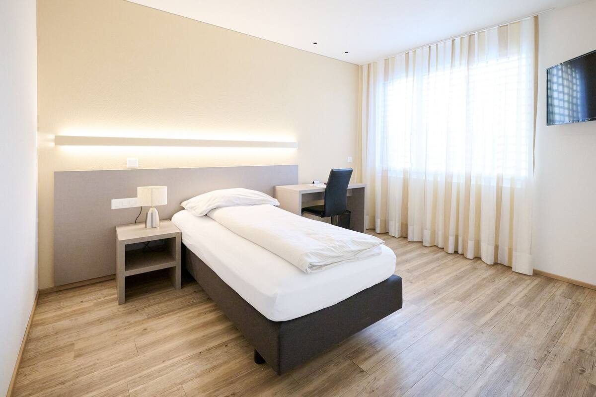 A modern hotel room with a bed, bedside table, lamp, desk, chair, and a TV mounted on the wall.