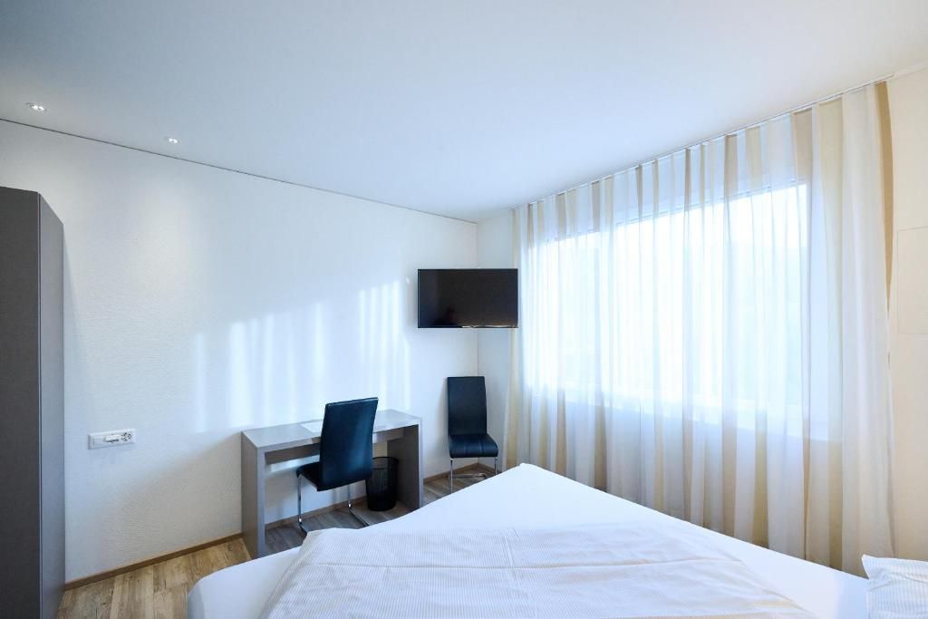 Quarto de hotel com cama, escrivaninha, cadeira, televisão e janela com cortinas.