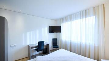 Quarto de hotel com cama, escrivaninha, cadeira, televisão e janela com cortinas.