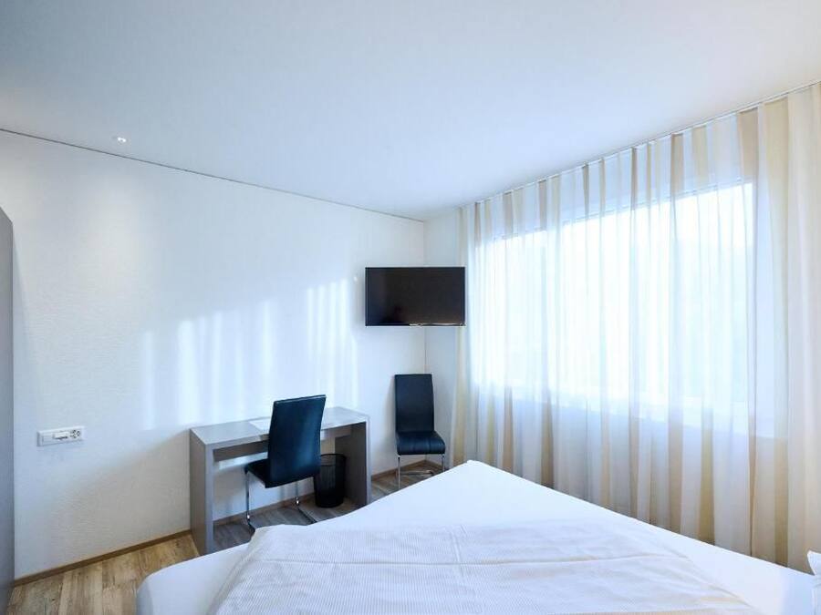 Quarto de hotel com cama, escrivaninha, cadeira, televisão e janela com cortinas.