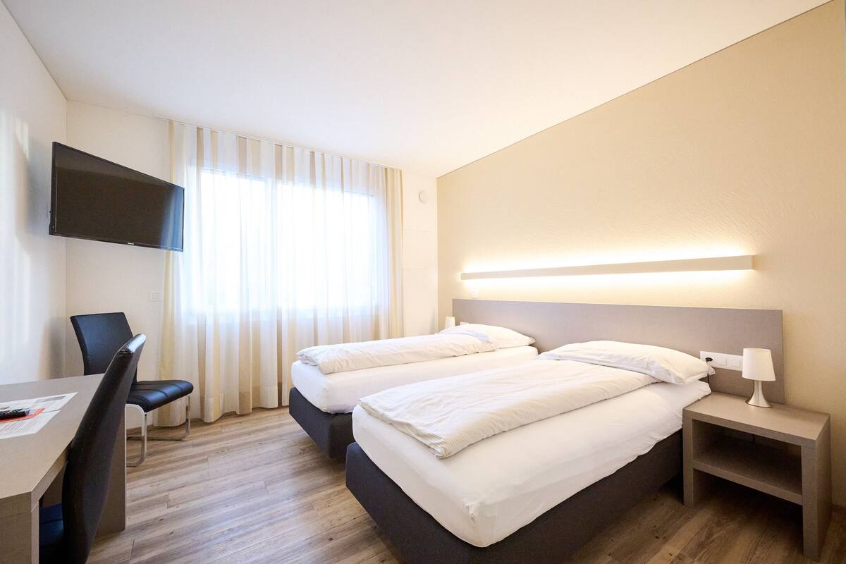 A hotel room with two beds, a desk, a chair, a TV, and a bedside table with a lamp.