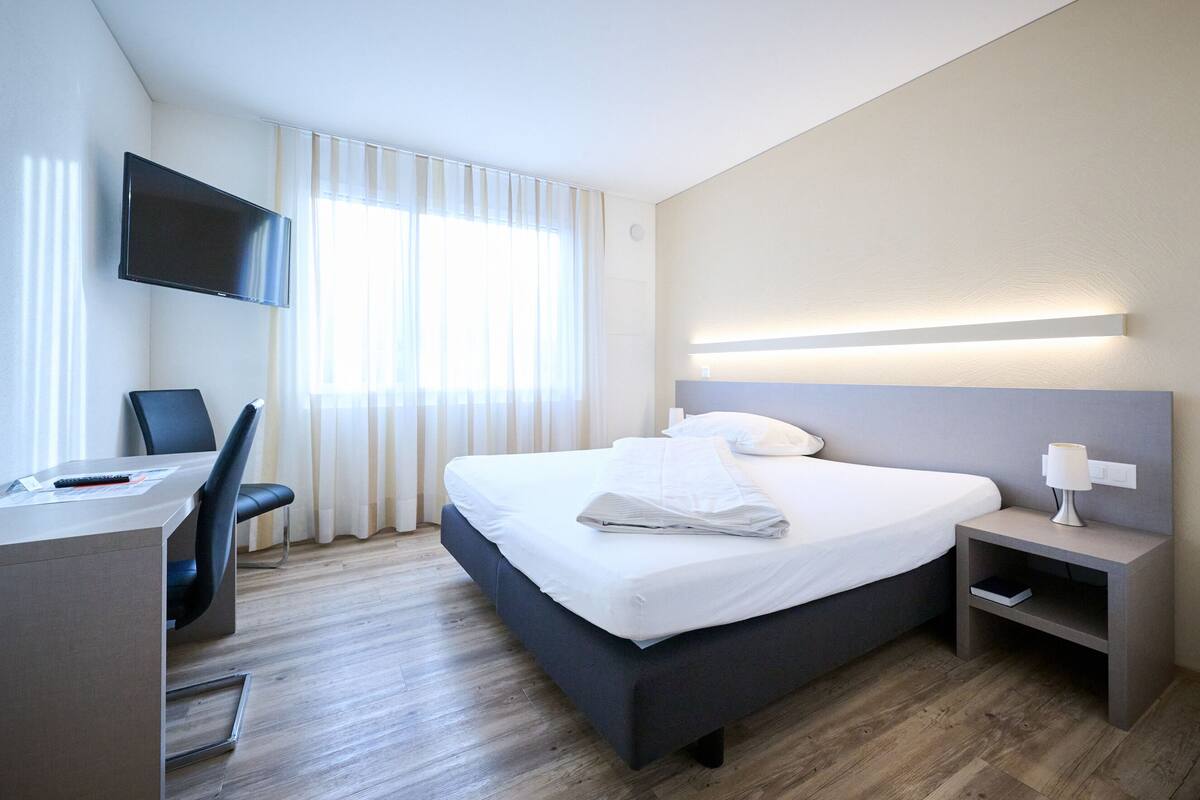 A hotel room with a bed, a desk, a chair, a TV mounted on the wall, and a bedside table with a lamp.