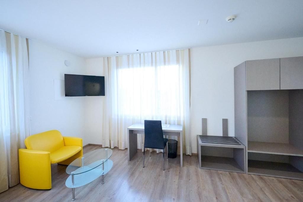 Ein modernes Hotelzimmer mit einem gelben Sessel, einem Glastisch, einem Schreibtisch mit Stuhl, einem flachbildfernseher an der Wand und einem eingebauten Kleiderschrank.