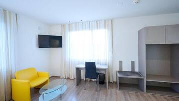 Ein modernes Hotelzimmer mit einem gelben Sessel, einem Glastisch, einem Schreibtisch mit Stuhl, einem flachbildfernseher an der Wand und einem eingebauten Kleiderschrank.