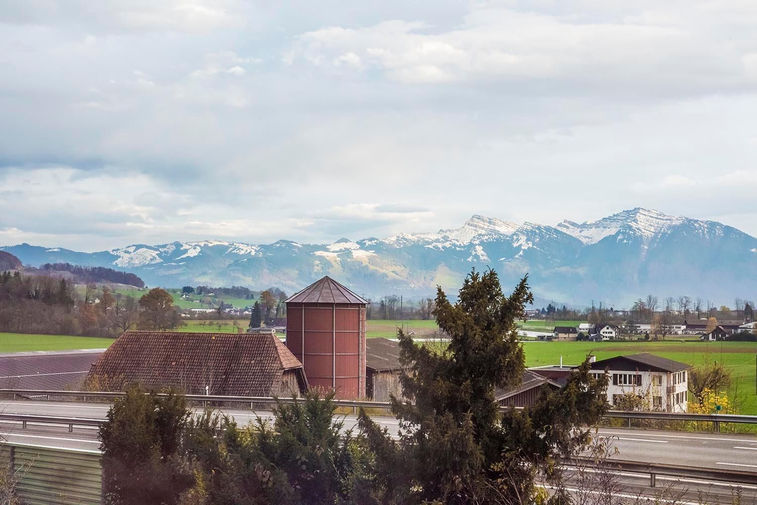 A rural landscape with a mountain range in the background, a silo, and a road in the foreground.