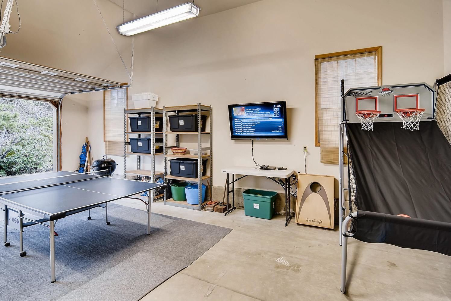 Un garage équipé d’une table de ping-pong, d’étagères avec des bacs de rangement, d’un téléviseur et d’un panier de basket.