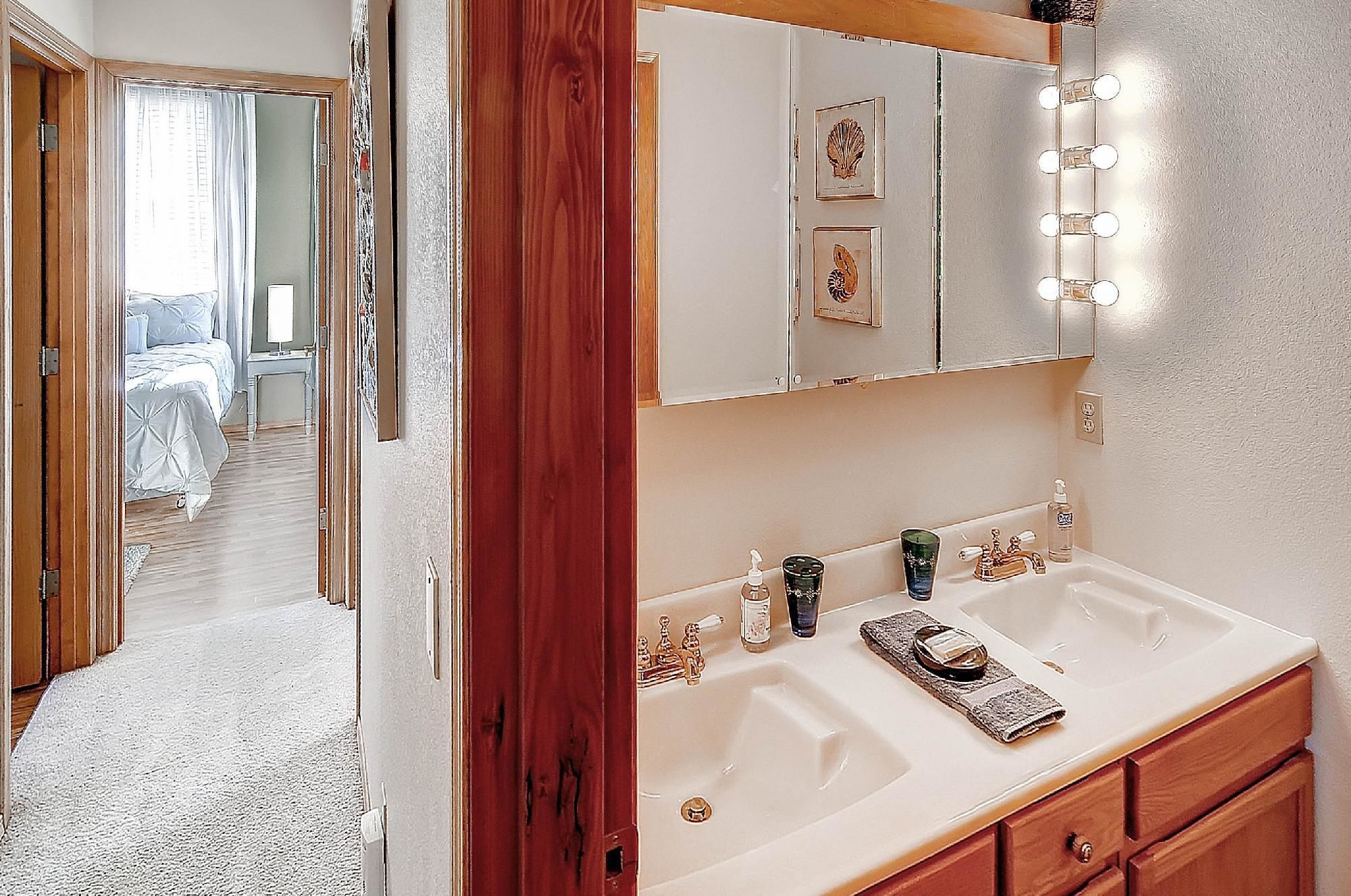 A bathroom with a vanity, sink, and mirror with lights. There is a bed visible in the adjacent room.