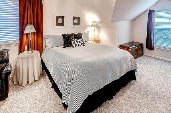 A bedroom with a large bed, two bedside lamps, a nightstand, a chest of drawers, and a window with blinds.