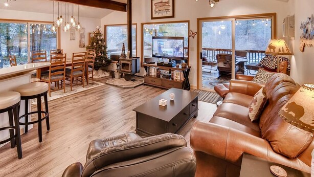 A cozy living room with a fireplace, leather sofa, wooden dining table, and large windows offering a view of the outdoors.