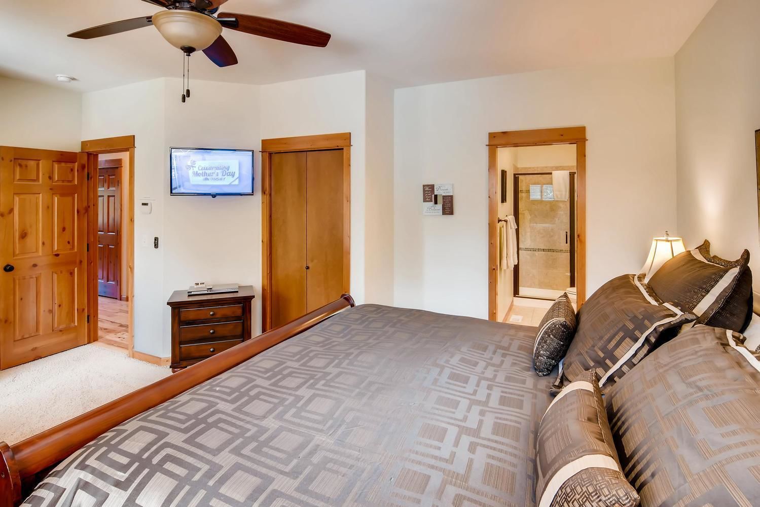 A bedroom with a large bed, a ceiling fan, a television, a wooden wardrobe, and a door leading to a bathroom.