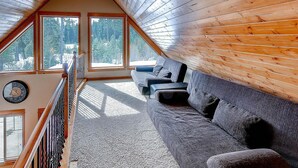 A cozy attic room with a wooden sloped ceiling, large windows, a carpeted floor, and a sofa with cushions. - Cascade Mountain Villa (Cle Elum)