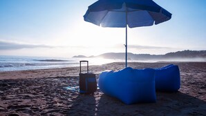 A beach scene with a blue umbrella, a blue bean bag, and a suitcase. - Casa Mono (Santa María Tonameca)