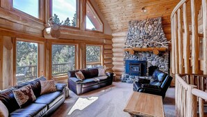 A log cabin interior with a stone fireplace, leather sofas, and large windows.