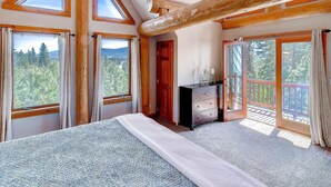 A bedroom with a large bed, wooden beams, and multiple windows offering views of a forested landscape.