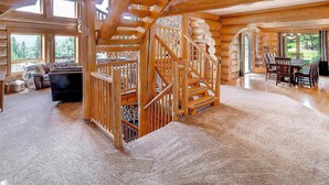 A spacious log cabin interior with a central wooden staircase, large windows, and a cozy seating area.
