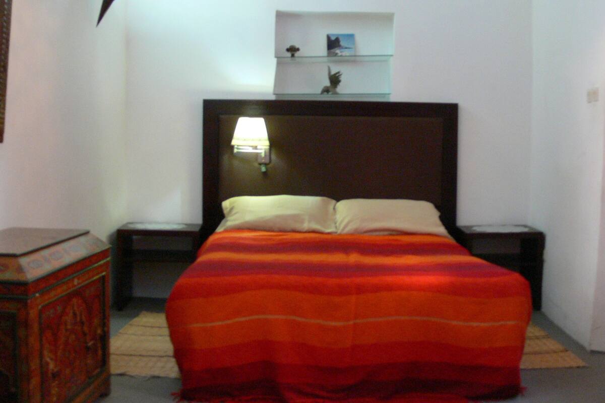 A bedroom with a bed, nightstands, a lamp, and a shelf with decorative items.