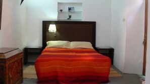 A bedroom with a bed, nightstands, a lamp, and a shelf with decorative items.