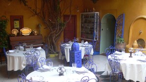 Un comedor con mesas redondas, sillas de metal azul y manteles blancos. Se observa un árbol y un aparador con puertas de vidrio al fondo.