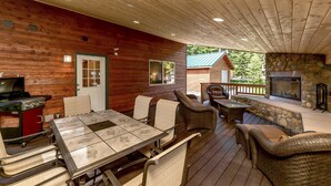 A wooden deck with a grill, a stone fireplace, wicker furniture, and a view of a cabin. - Tall Pines (Cle Elum)