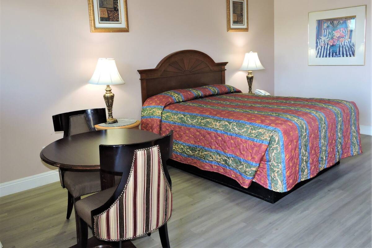 A hotel room with a bed, two bedside lamps, a round table with a chair, and two framed artworks on the walls.