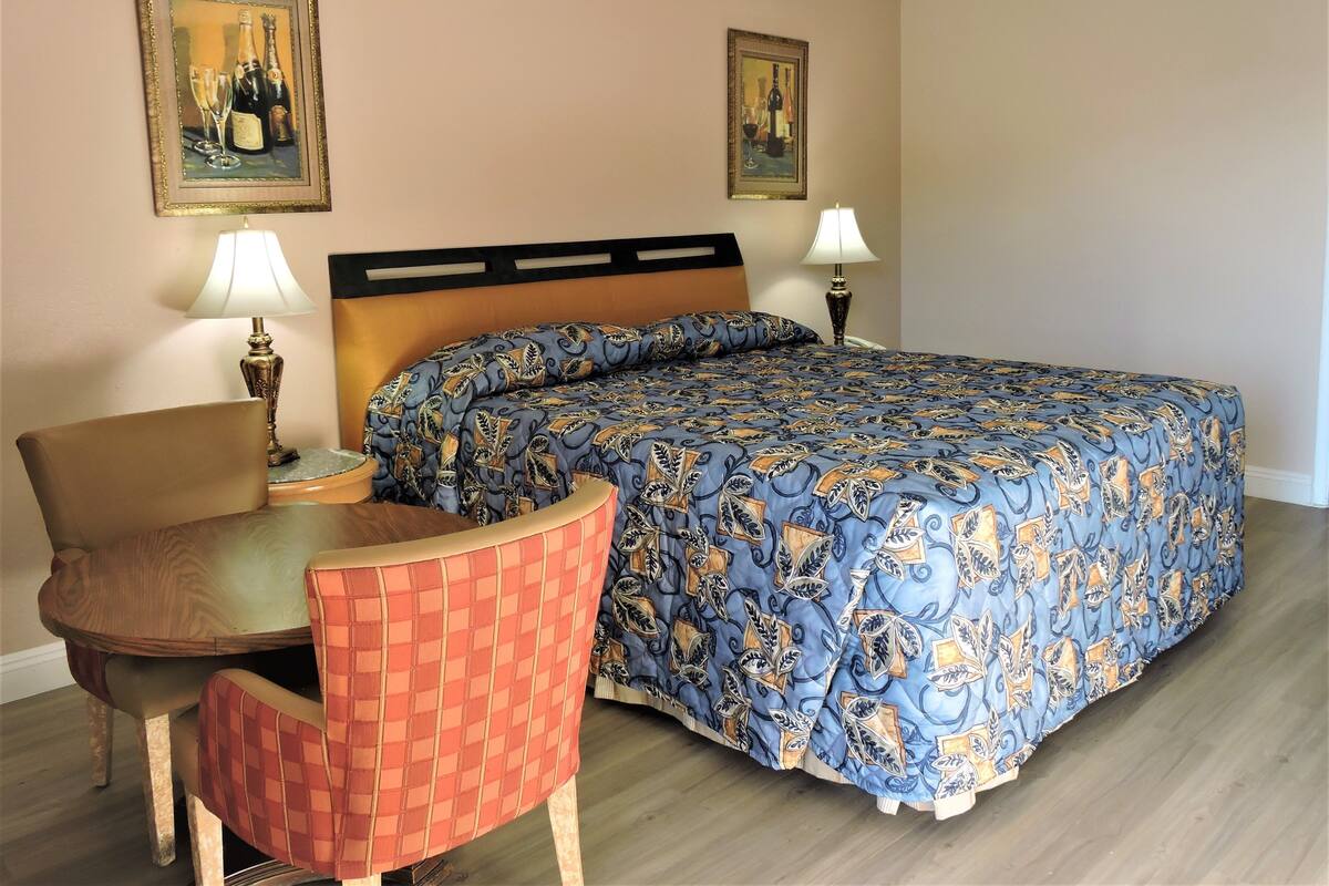 A hotel room with a bed, a chair, a table, two lamps, and two framed pictures on the wall.