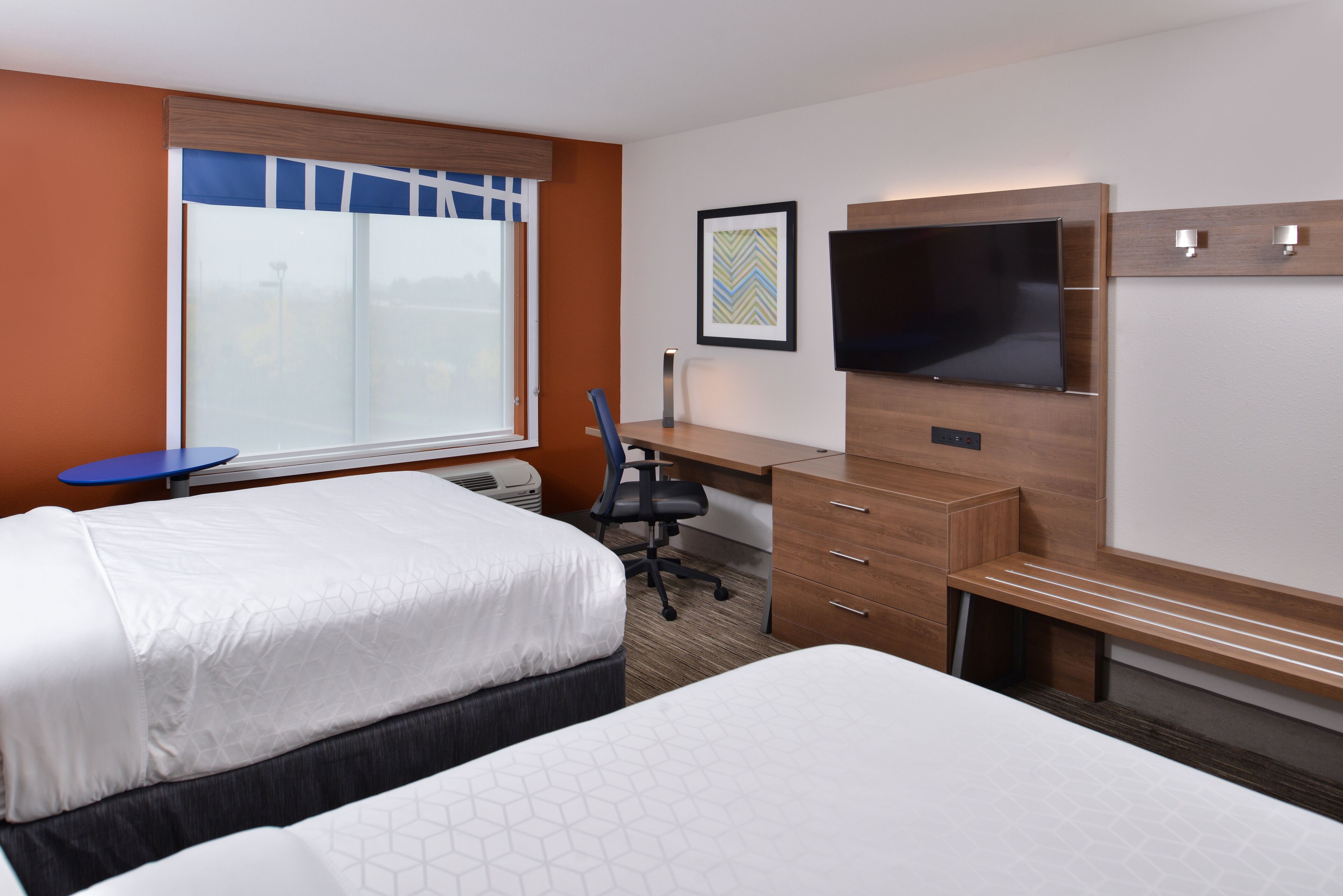 A hotel room with two beds, a desk, a chair, a TV, and a window with blinds.