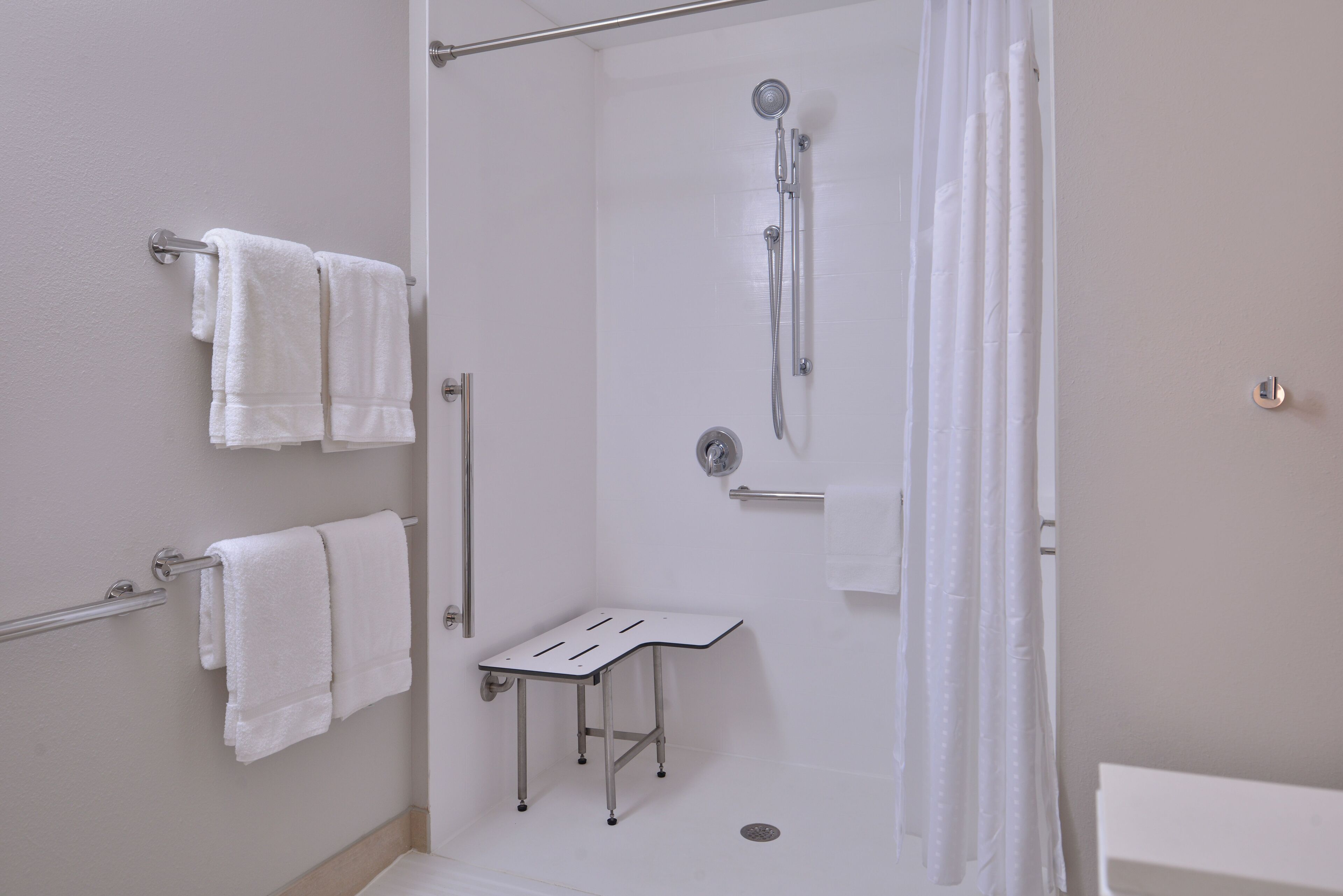 Une douche à l’italienne avec un pommeau de douche amovible, un petit banc blanc et deux porte-serviettes avec des serviettes blanches.