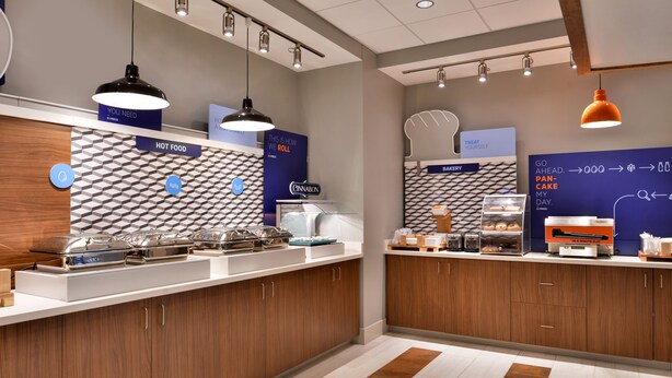 A modern breakfast area with a counter displaying various food items, a bakery section, and a sign indicating the need for a pan. - Holiday Inn Express & Suites Marshalltown by IHG (Marshalltown)