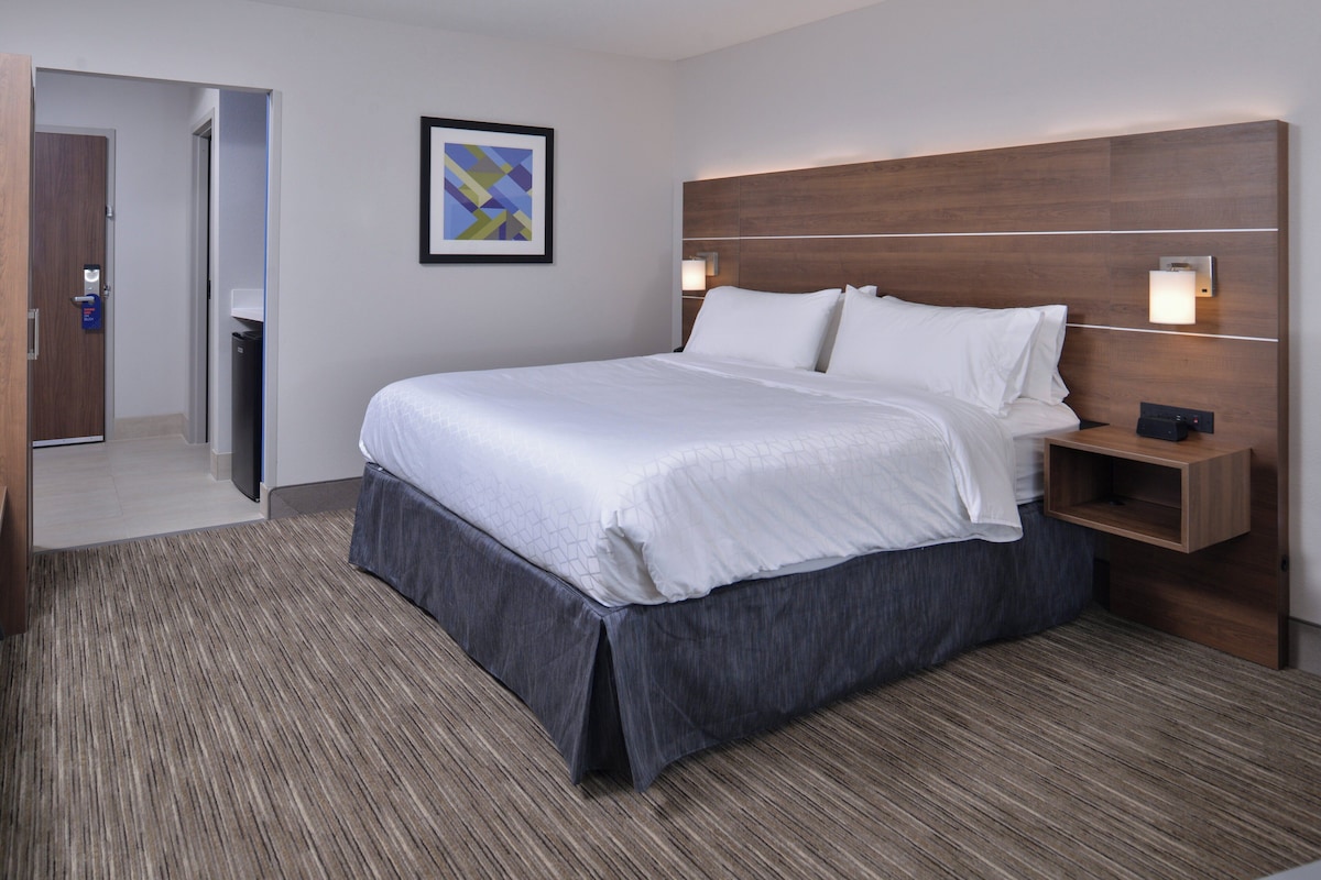 Habitación de hotel con una cama grande, una mesita de noche, una lámpara de pared y una puerta que da a otra habitación.