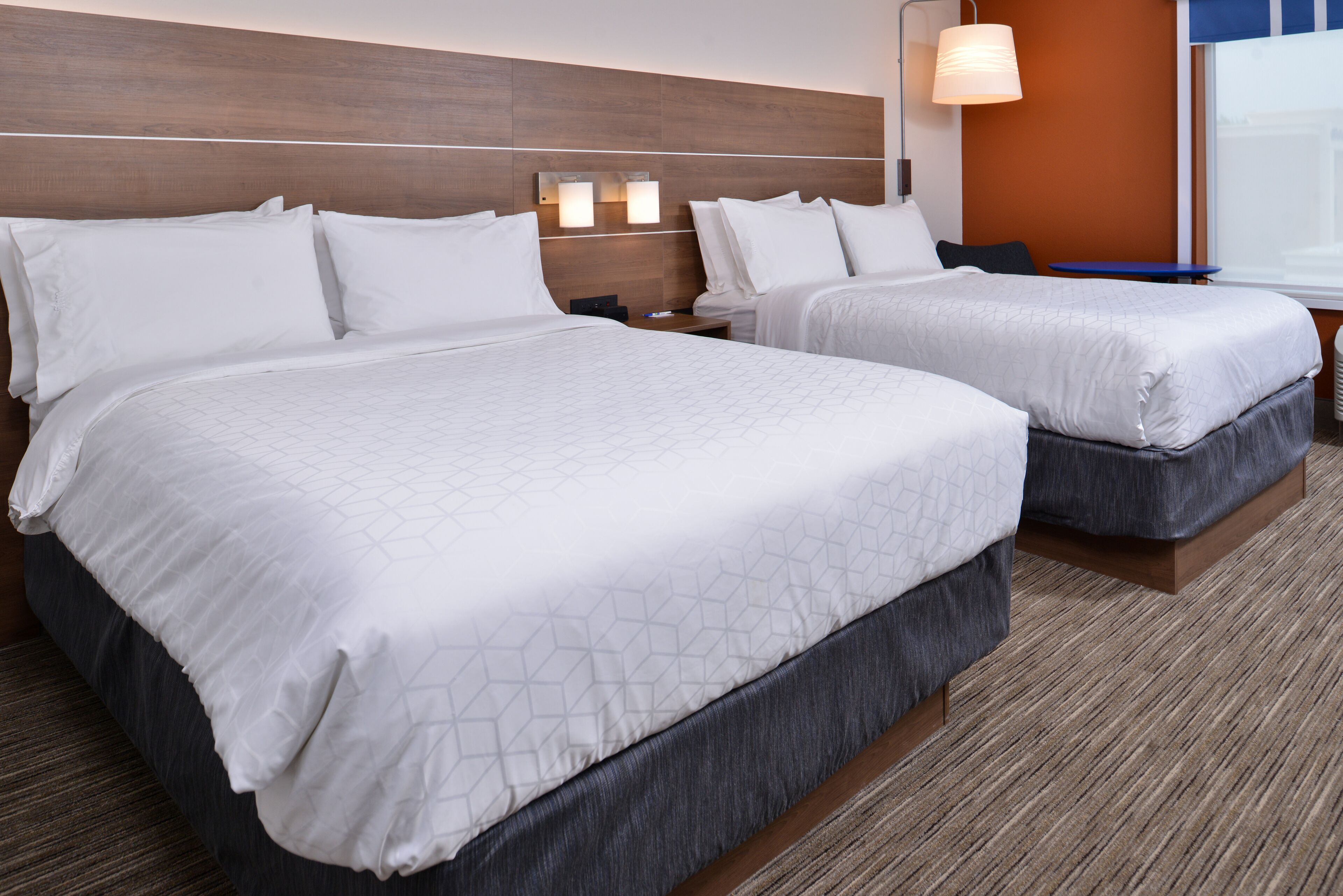 A hotel room with two beds, a wooden headboard, a nightstand, and a wall-mounted lamp.