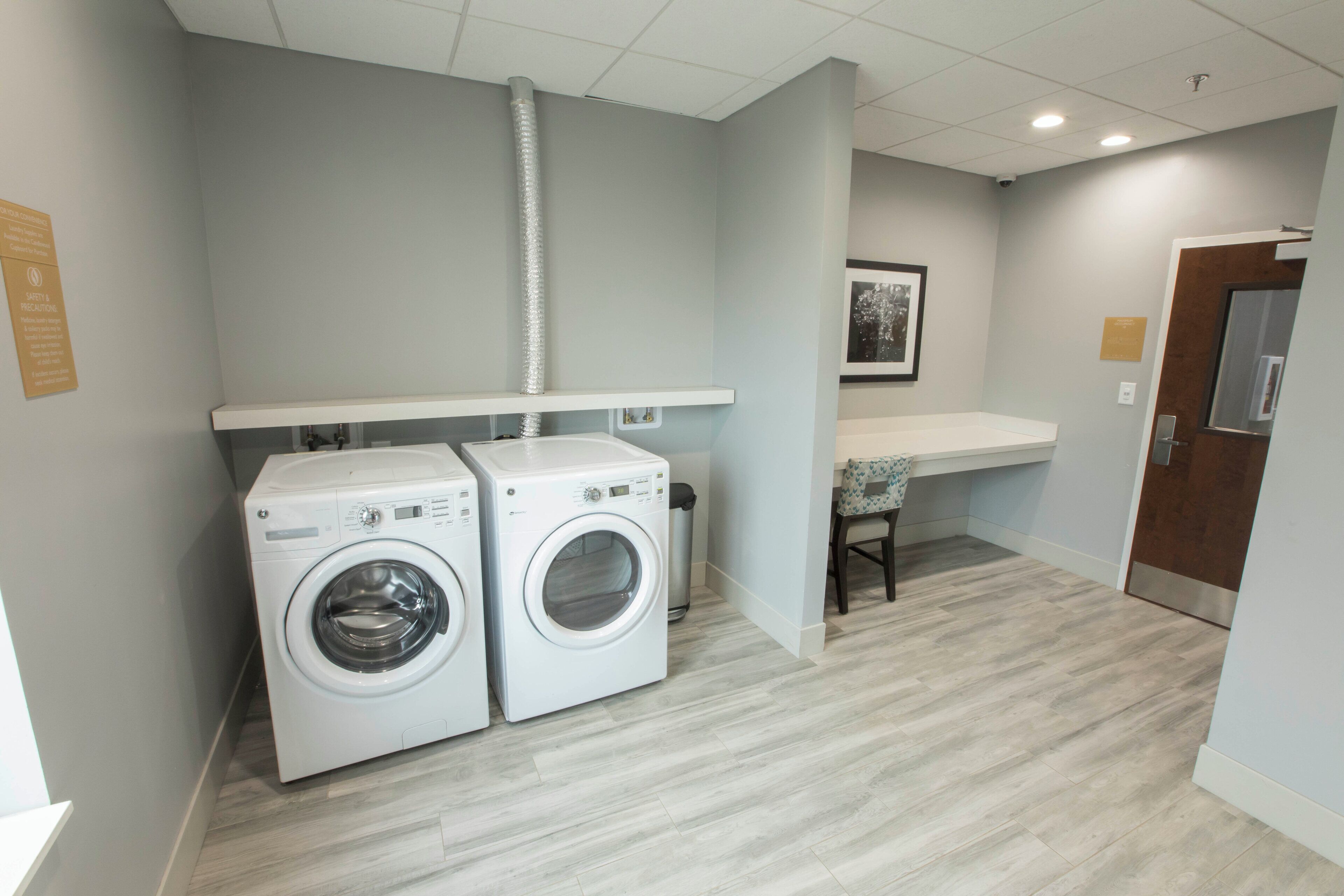 A laundry room with two washing machines, a ventilation duct, a trash bin, and a small seating area with a chair.