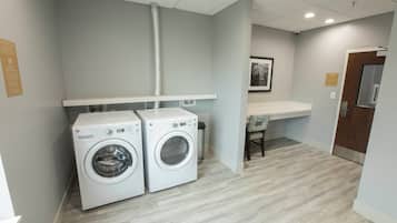 A laundry room with two washing machines, a ventilation duct, a trash bin, and a small seating area with a chair.