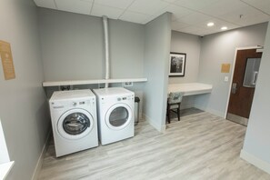 A laundry room with two washing machines, a ventilation duct, a trash bin, and a small seating area with a chair. - Candlewood Suites McDonough by IHG (McDonough)