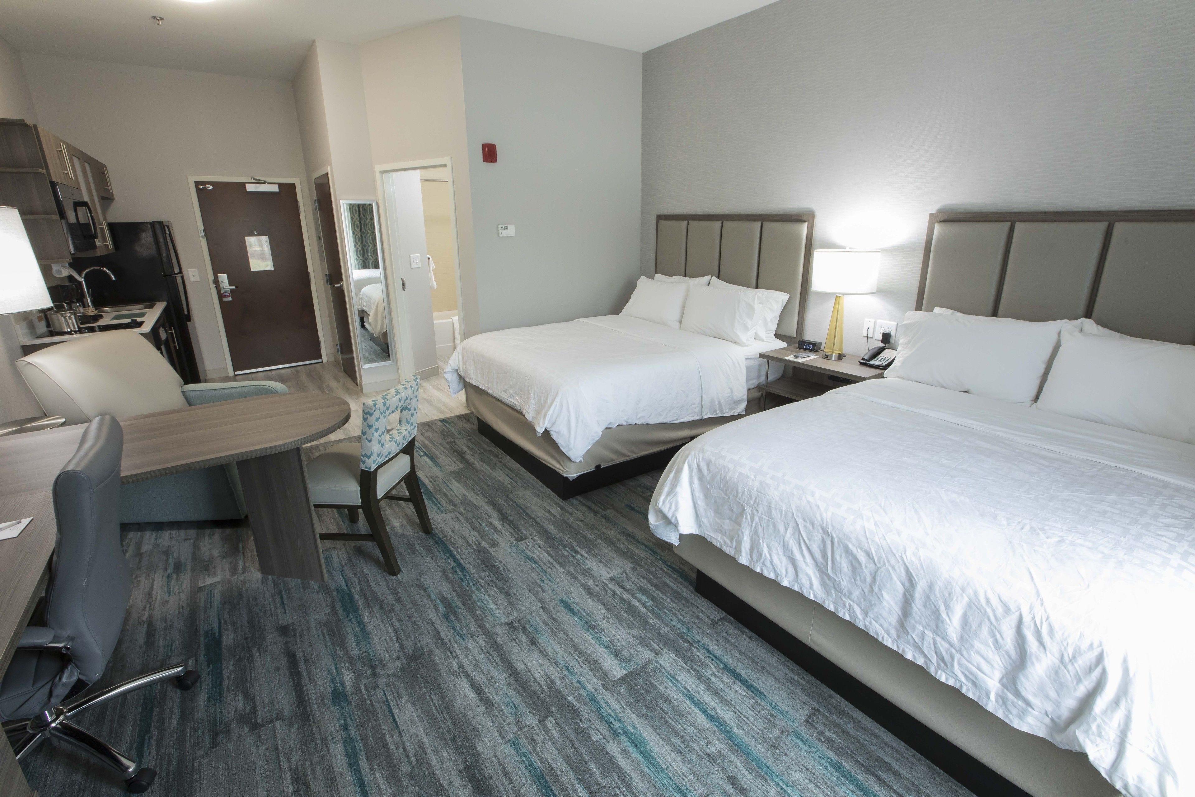 A hotel room with two beds, a desk, a chair, a small kitchenette, and a bathroom door.