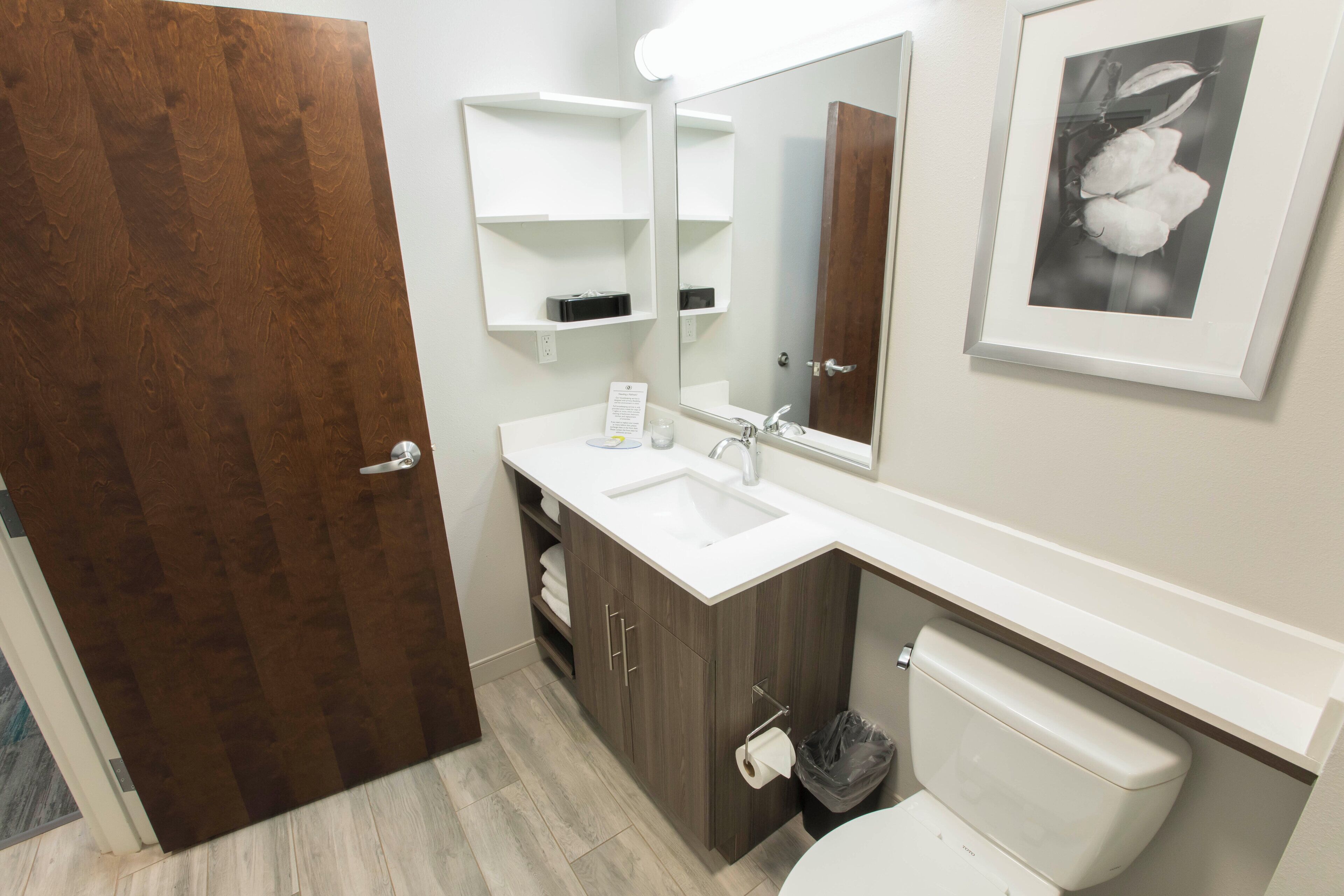 A bathroom with a wooden door, a white sink, a mirror, a toilet, and a framed picture on the wall.