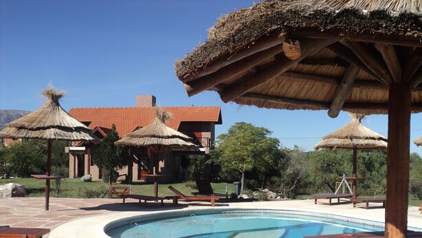 A swimming pool with lounge chairs and thatched umbrellas, surrounded by a stone patio and a building with a red-tiled roof. - Cabañas Oma Lissi (Merlo)