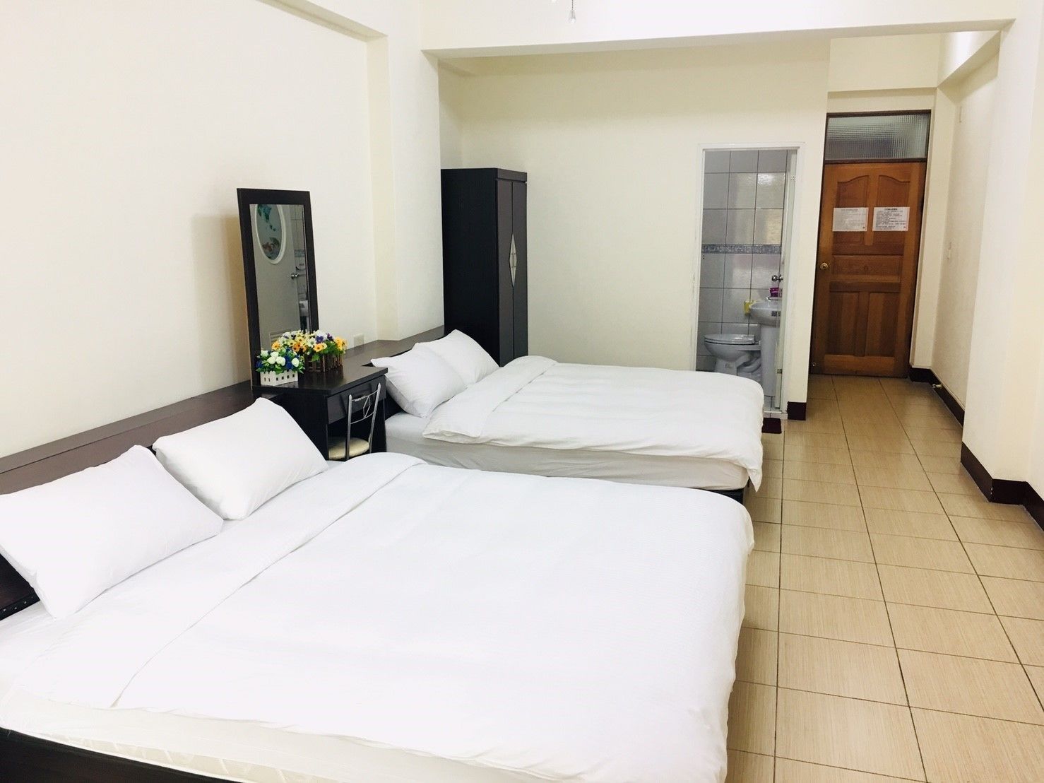 A hotel room with two beds, a desk with a mirror, and a small table with flowers.