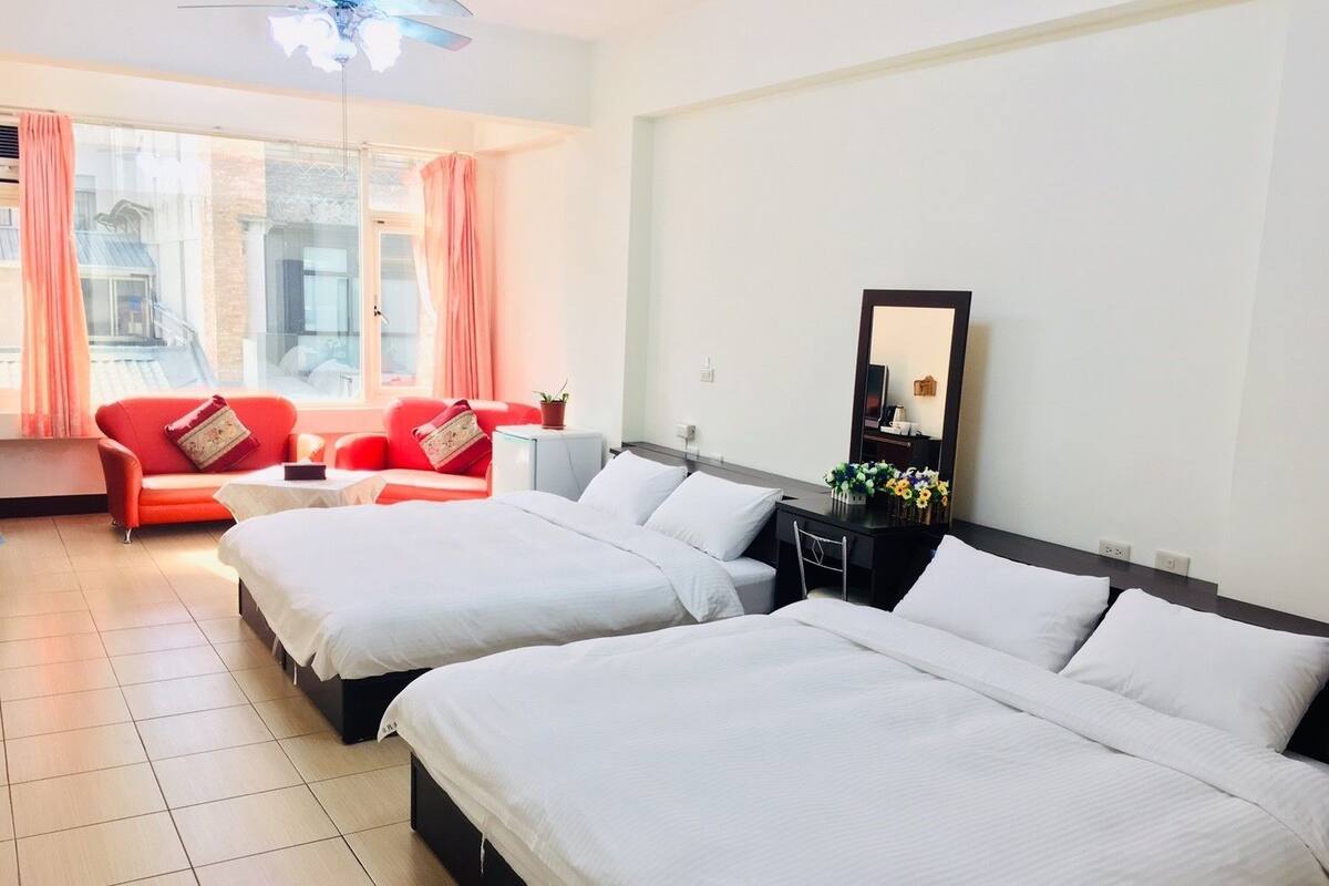 Habitación de hotel con dos camas, un sofá rojo, una mesita y un escritorio con espejo y flores.