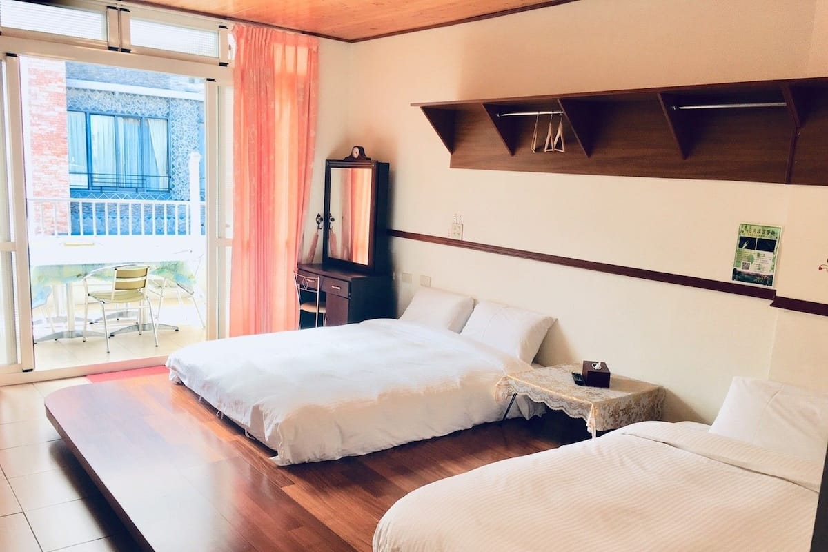 Habitación de hotel con dos camas, suelo de madera, un balcón con una silla y un estante de madera sobre las camas.