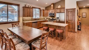 A modern kitchen with wooden cabinets, a central island with stools, stainless steel appliances, and a large window with blinds. - Lake Cabins Lodge (Cle Elum)