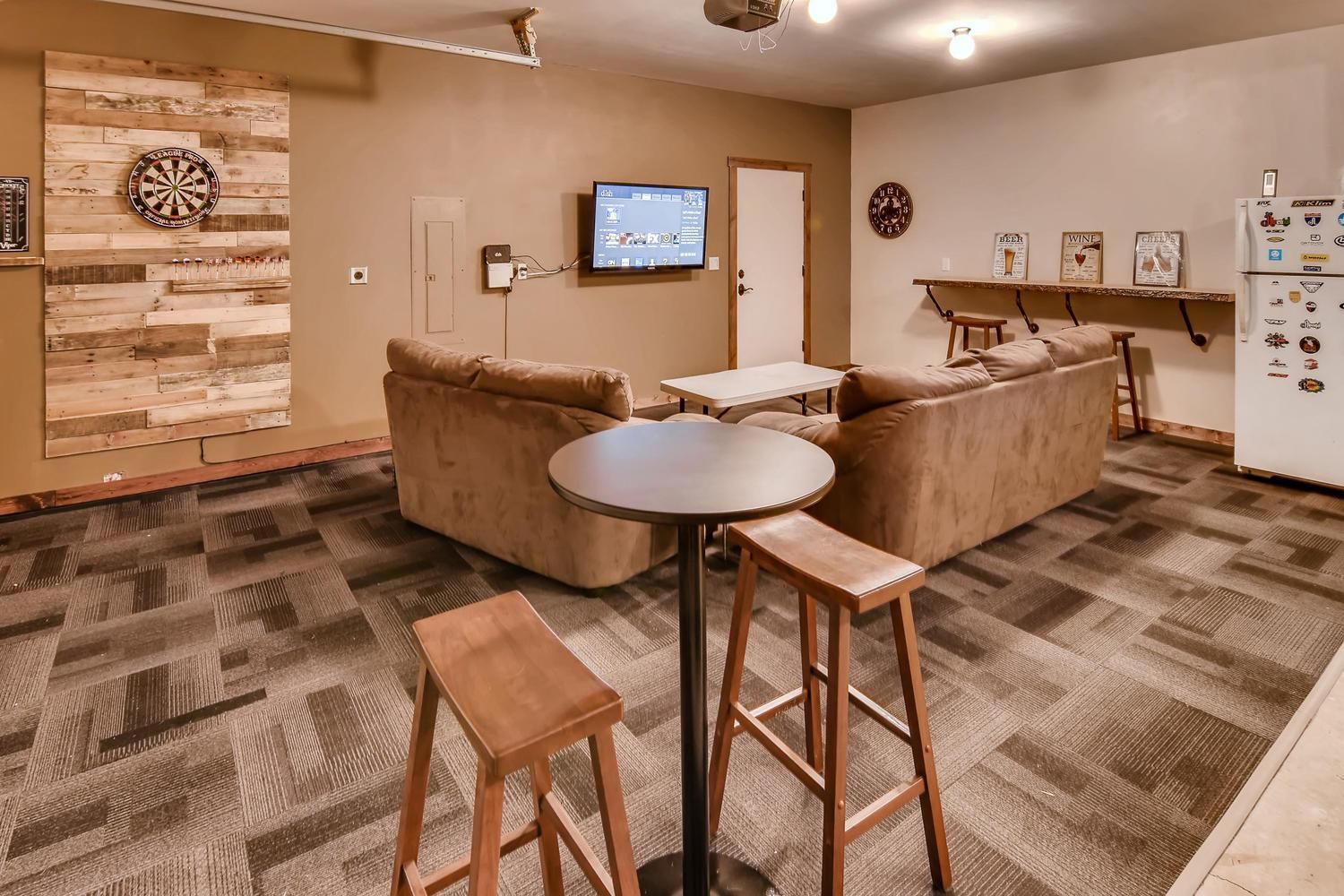 Un salon chaleureux avec un mur en bois, un jeu de fléchettes, un téléviseur et un réfrigérateur décoré d’aimants.