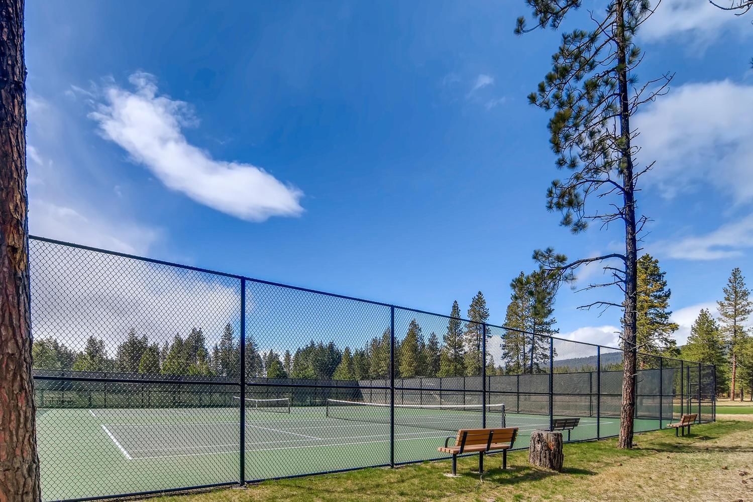 A tennis court enclosed by a chain-link fence with benches and trees in the background.