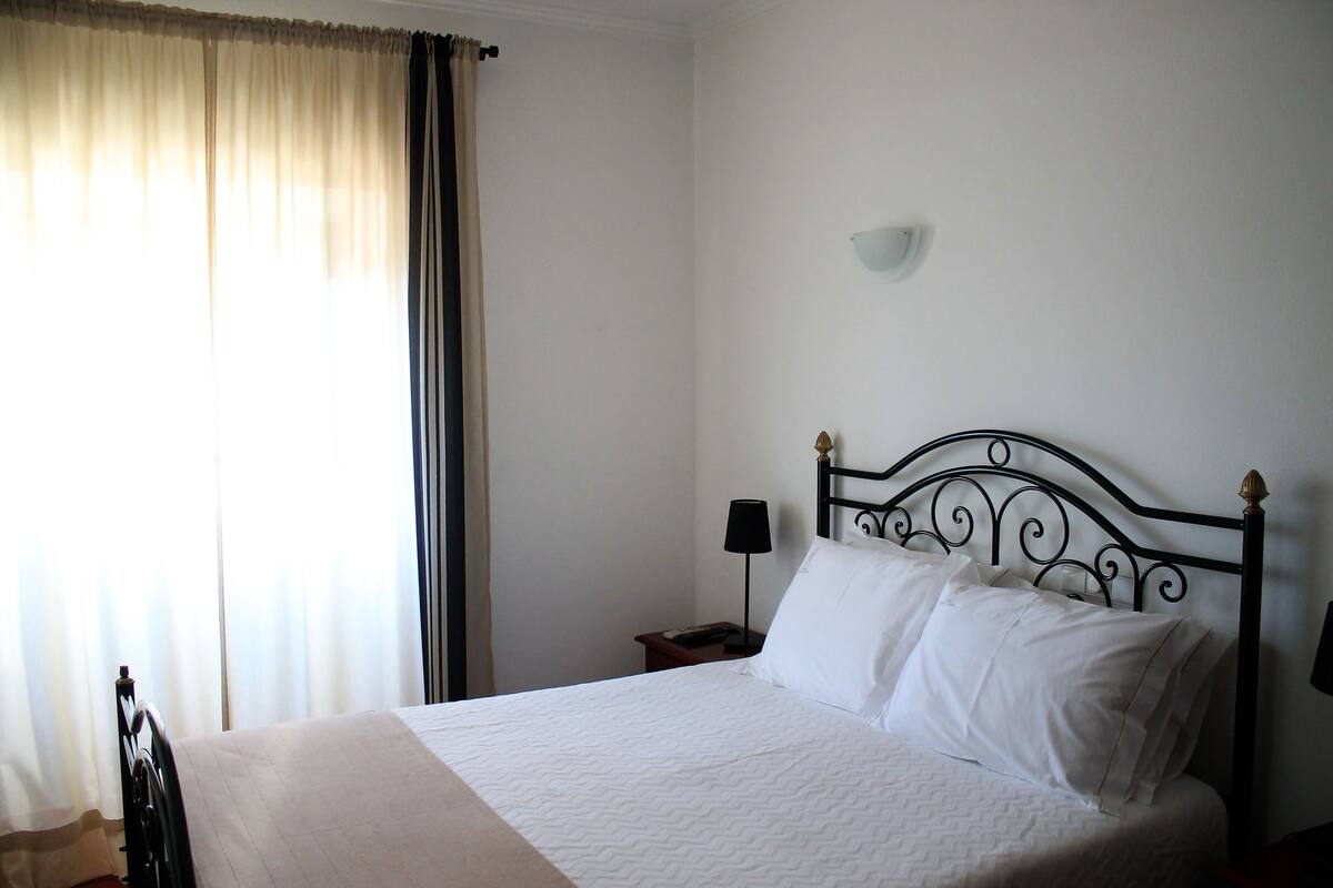 Una cama bien hecha con sábanas blancas, cabecera de metal negro y una mesita de noche con lámpara.