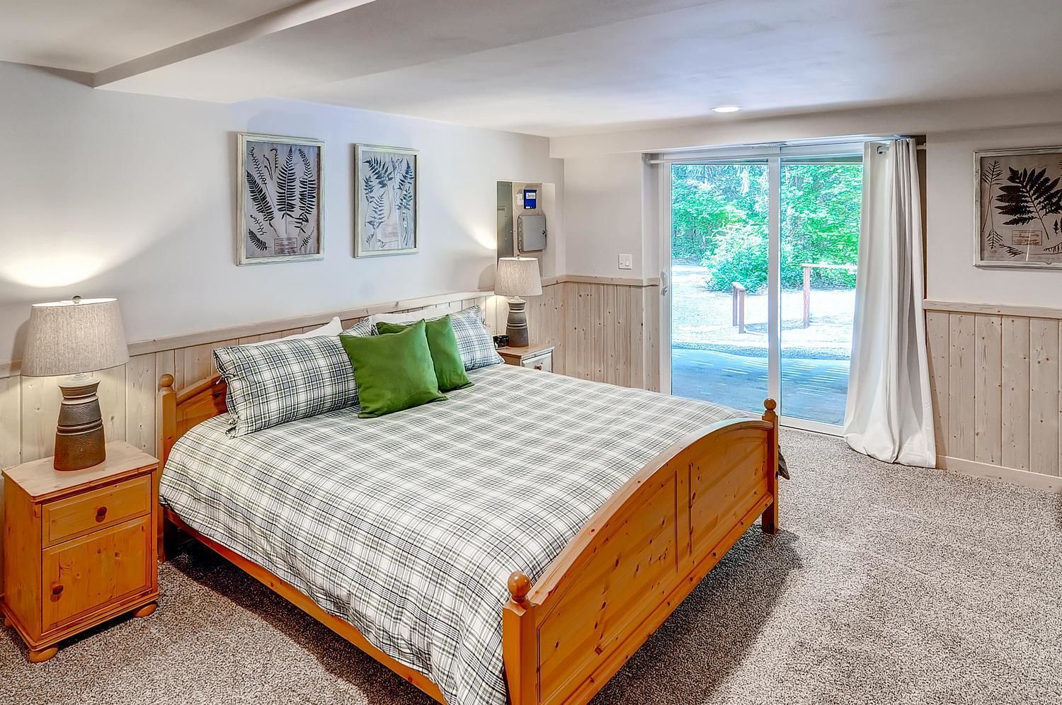 A bedroom with a wooden bed, two bedside tables, a nightstand, and a large window with a view of greenery.