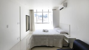 Ein minimalistisch eingerichtetes Hotelzimmer mit einem großen Fenster, einem Bett mit weißer Bettdecke, einem Nachttisch und einem schwarzen Stuhl.