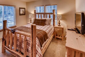 A bedroom with a wooden bed frame, a plaid bedspread, two bedside tables with lamps, a television, and windows with blinds. - Roslyn Chalet (Cle Elum)