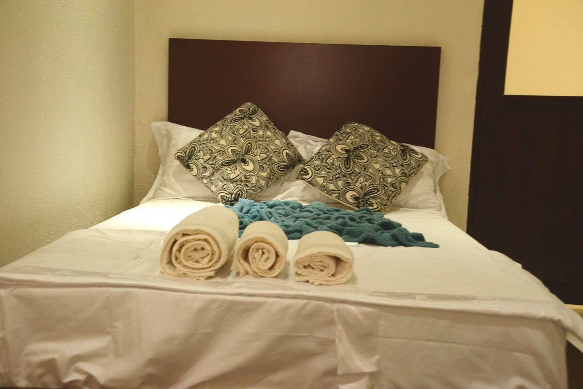 A neatly made bed with patterned pillows, a blue throw blanket, and rolled towels.